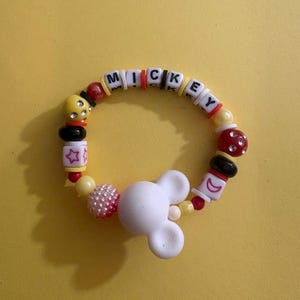 Op de afbeelding: Een kleurrijke kralenarmband met de naam "MICKEY" gespeld in witte letterkralen. De armband heeft een witte Mickey Mouse-hoofdbedeltje, rode, gele, zwarte en roze kralen. Op een gele achtergrond.