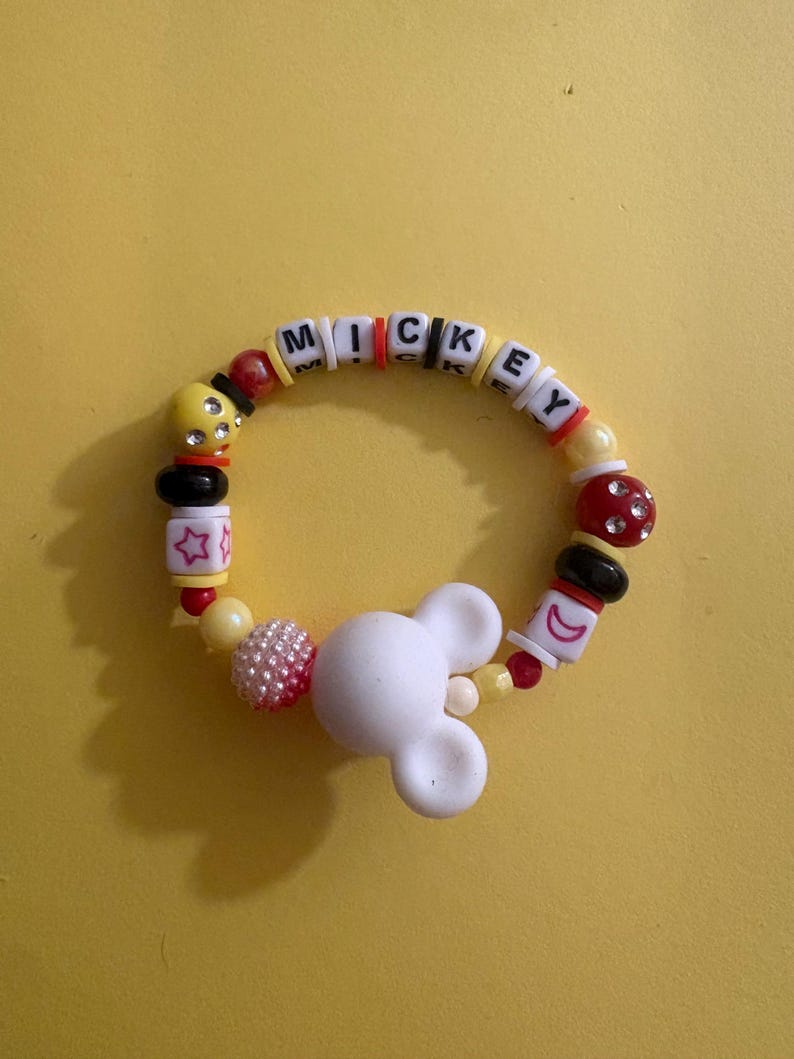 Op de afbeelding: Een kleurrijke kralenarmband met de naam "MICKEY" gespeld in witte letterkralen. De armband heeft een witte Mickey Mouse-hoofdbedeltje, rode, gele, zwarte en roze kralen. Op een gele achtergrond.
