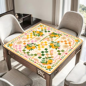 May include: A square table cover with a colorful citrus fruit design. The cover features oranges, orange slices, and floral accents on a cream background with a checkered pattern. The border is adorned with pink flowers and green leaves.