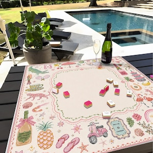 Pu&ograve; includere: Un tappetino da mahjong quadrato con un centro color crema e un bordo smerlato rosa. Il bordo presenta illustrazioni colorate di uno scooter rosa, bottiglie di champagne, un ananas, infradito, un leopardo, un fenicottero e una pagoda. Diverse tessere del mahjong sono sparse sul tappetino, posizionato su un tavolo scuro vicino a una piscina.