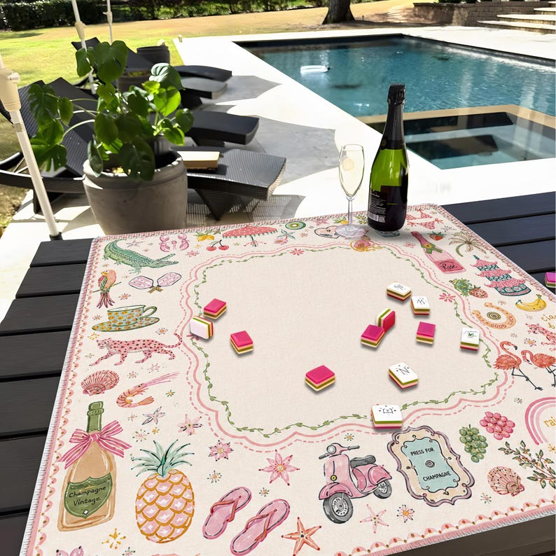 Pu&ograve; includere: Un tappetino da mahjong quadrato con un centro color crema e un bordo smerlato rosa. Il bordo presenta illustrazioni colorate di uno scooter rosa, bottiglie di champagne, un ananas, infradito, un leopardo, un fenicottero e una pagoda. Diverse tessere del mahjong sono sparse sul tappetino, posizionato su un tavolo scuro vicino a una piscina.