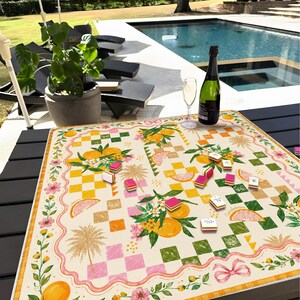 May include: A decorative table setting with a colorful, patterned tablecloth featuring oranges, flowers, and palm trees. The table is set with a bottle of wine, a glass, and game pieces. The scene is outdoors near a pool.