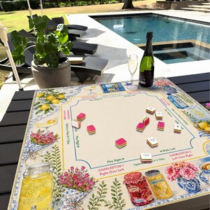 May include: A decorative game board with a floral and lemon design, featuring pink and white game tiles. A bottle of champagne and a glass are on the table. The board has text that says "Charleston".