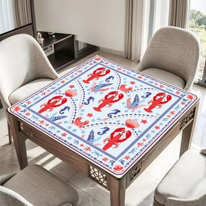K&ouml;nnte beinhalten: Eine quadratische Tischdecke mit einem wiederkehrenden Muster aus roten Hummern, blauen Seepferdchen und korallenfarbenen floralen Elementen auf wei&szlig;em Grund. Der Rand zeigt ein blaues geometrisches M&auml;ander-Muster mit kleinen roten Sternen.