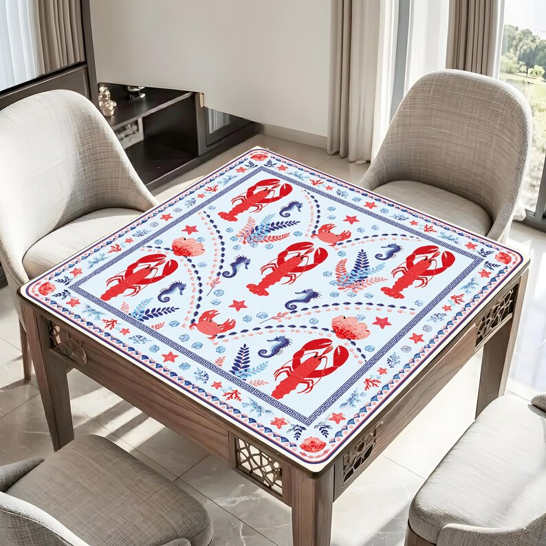 K&ouml;nnte beinhalten: Eine quadratische Tischdecke mit einem wiederkehrenden Muster aus roten Hummern, blauen Seepferdchen und korallenfarbenen floralen Elementen auf wei&szlig;em Grund. Der Rand zeigt ein blaues geometrisches M&auml;ander-Muster mit kleinen roten Sternen.