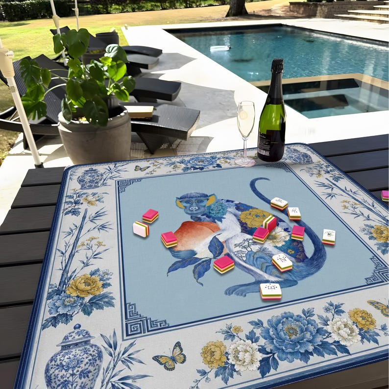 K&ouml;nnte beinhalten: Blau-wei&szlig;e dekorative Mahjong-Spielmatte mit einer Affenillustration und einer Pfirsich. Die Matte hat einen floralen Rand mit Schmetterlingen und einem Vasenmuster. Eine Flasche Champagner und ein Glas stehen auf dem Tisch.