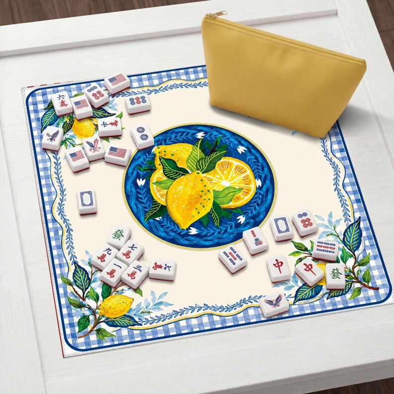 May include: A yellow zippered pouch rests on a blue and white patterned mat with a lemon and leaf design. Scattered around are white mahjong tiles, some with American flag designs and Chinese characters.