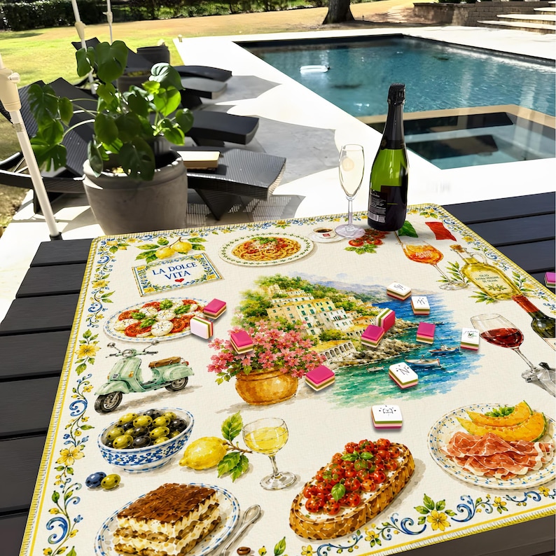 May include: A square tablecloth featuring a colorful Italian-themed print with illustrations of tiramisu, bruschetta, olives, a scooter, and a coastal village. The text LA DOLCE VITA is printed on the fabric, which is scattered with small game tiles.