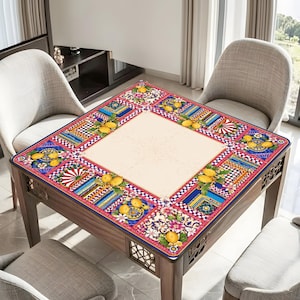 Pode incluir: Mesa quadrada de madeira com um tampo estampado colorido. A borda apresenta desenhos florais, geom&eacute;tricos e de frutas em vermelho, azul, amarelo e branco. A mesa &eacute; cercada por quatro cadeiras cinzentas. Aproximadamente 90 cm x 90 cm.