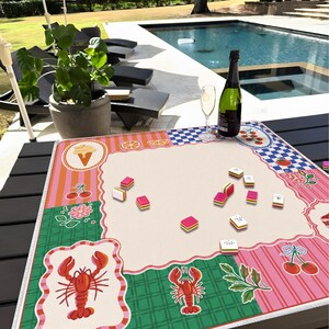 May include: A colorful mahjong game mat with a cream-colored center and decorative borders featuring ice cream cones, cherries, and lobsters. A bottle of champagne and a glass are on the table, along with mahjong tiles.