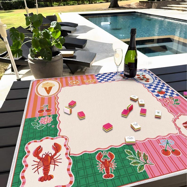 May include: A colorful mahjong game mat with a cream-colored center and decorative borders featuring ice cream cones, cherries, and lobsters. A bottle of champagne and a glass are on the table, along with mahjong tiles.
