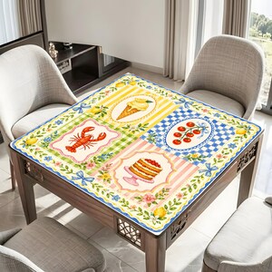 May include: A square table with a decorative tablecloth featuring food illustrations. The tablecloth has a lobster, ice cream cone, cake, and cherry design. The table is surrounded by chairs and has a wooden frame.