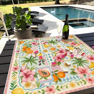 Pu&ograve; includere: Un colorato tabellone di gioco con motivi floreali, frutta tropicale, palme e fiori rosa. Il tabellone &egrave; appoggiato su un tavolo scuro, con una bottiglia di champagne e un bicchiere nelle vicinanze. Sullo sfondo una piscina e sedie a sdraio.
