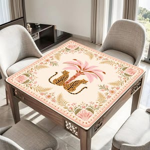 May include: A square table cover featuring a cream-colored background with a central illustration of two leopards resting beneath a pink palm tree. The border is decorated with a symmetrical pattern of pink flowers, green ferns, and fan-shaped motifs.
