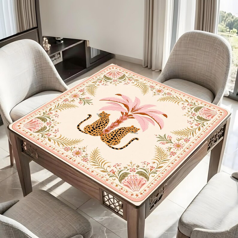 May include: A square table cover featuring a cream-colored background with a central illustration of two leopards resting beneath a pink palm tree. The border is decorated with a symmetrical pattern of pink flowers, green ferns, and fan-shaped motifs.