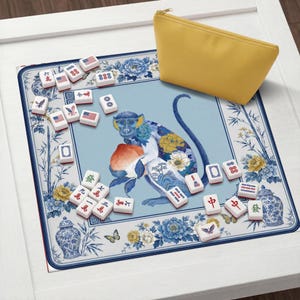 K&ouml;nnte beinhalten: Ein Mahjong-Set mit Spielsteinen und einem gelben Rei&szlig;verschlussetui. Die Spielmatte zeigt eine blaue Affenillustration, die einen Pfirsich h&auml;lt, umgeben von Blumen- und Bambusdesigns. Die Spielsteine haben verschiedene Symbole und amerikanische Flaggen.