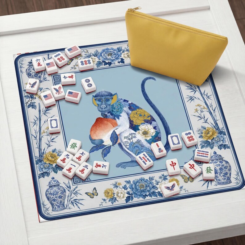 K&ouml;nnte beinhalten: Ein Mahjong-Set mit Spielsteinen und einem gelben Rei&szlig;verschlussetui. Die Spielmatte zeigt eine blaue Affenillustration, die einen Pfirsich h&auml;lt, umgeben von Blumen- und Bambusdesigns. Die Spielsteine haben verschiedene Symbole und amerikanische Flaggen.