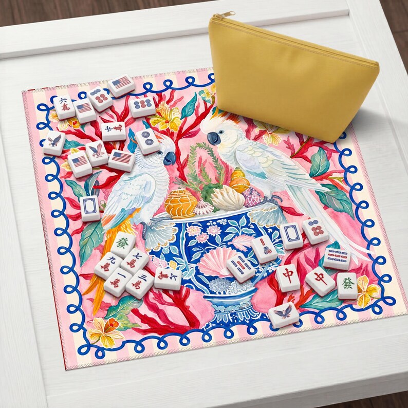 May include: A decorative Mahjong set with white tiles featuring Chinese characters and American flag designs. The set is arranged on a pink floral mat with two white cockatoos and a yellow zippered pouch.