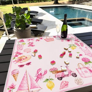 Puede incluir: Una alfombrilla de tela cuadrada de color rosa con un patr&oacute;n veraniego de veleros, langostas, limones, conos de helado, scooters, bolsas de playa, conchas y flores, con varias fichas de juego rectangulares dispersas por la superficie.