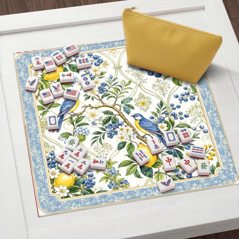 K&ouml;nnte beinhalten: Eine quadratische Mahjong-Matte mit wei&szlig;em Hintergrund, blauen V&ouml;geln, gelben Zitronen und blauen Beeren, umrahmt von einer blauen Blumenbord&uuml;re. Verstreute wei&szlig;e Mahjong-Steine mit verschiedenen Symbolen, US-Flaggen und V&ouml;geln liegen auf der Matte, dazu eine gelbe Tasche.