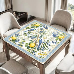 K&ouml;nnte beinhalten: Eine quadratische Tischdecke mit wei&szlig;em Hintergrund und einem botanischen Muster aus gelben Zitronen, blauen V&ouml;geln und dunkelblauen Beerenclustern. Das Design ist von einem dekorativen hellblauen Rand mit dezentem Blumenmuster umrahmt.