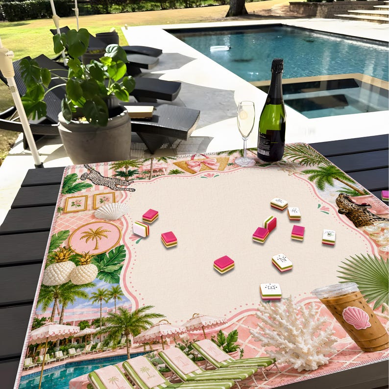 May include: A pink and cream-colored mahjong game set on a table with a tropical theme. The set includes tiles, a decorative mat with palm trees, pineapples, and a pool scene, a bottle of champagne, and a glass of champagne.