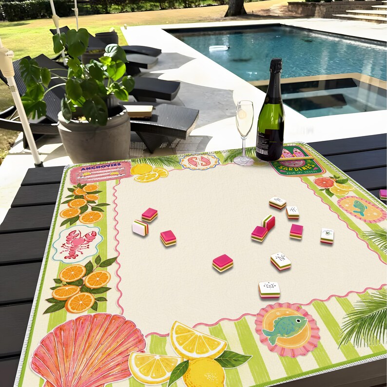 May include: A table setting featuring a decorative mat with fruit and seafood illustrations, including oranges, lemons, and a lobster. Scattered game pieces are on the mat. A bottle of champagne and a glass are also present.