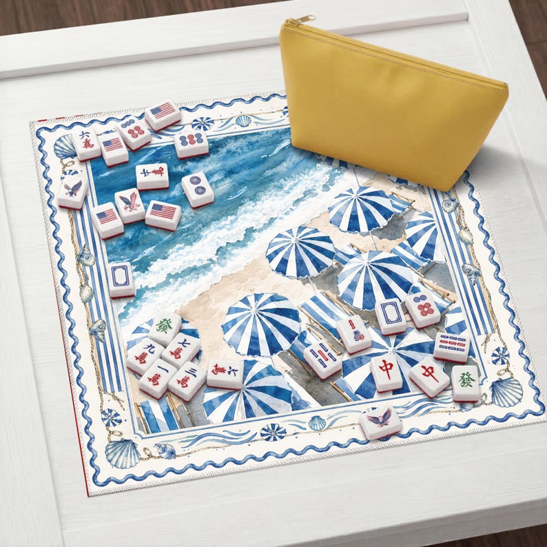 Op de afbeelding: Een vierkante mahjongmat met een aquarel strandsc&egrave;ne met blauw-wit gestreepte parasols, verspreide mahjongtegels met Amerikaanse vlaggen en traditionele symbolen, en een effen geel etui met ritssluiting in de hoek.