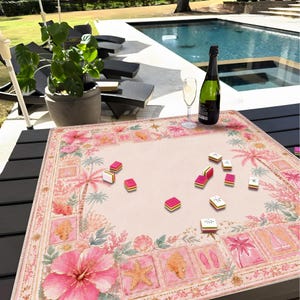 May include: A square game mat featuring a light pink center and a decorative border with illustrations of pink hibiscus flowers, seashells, starfish, palm trees, and sailboats, with several small rectangular game tiles scattered on top.