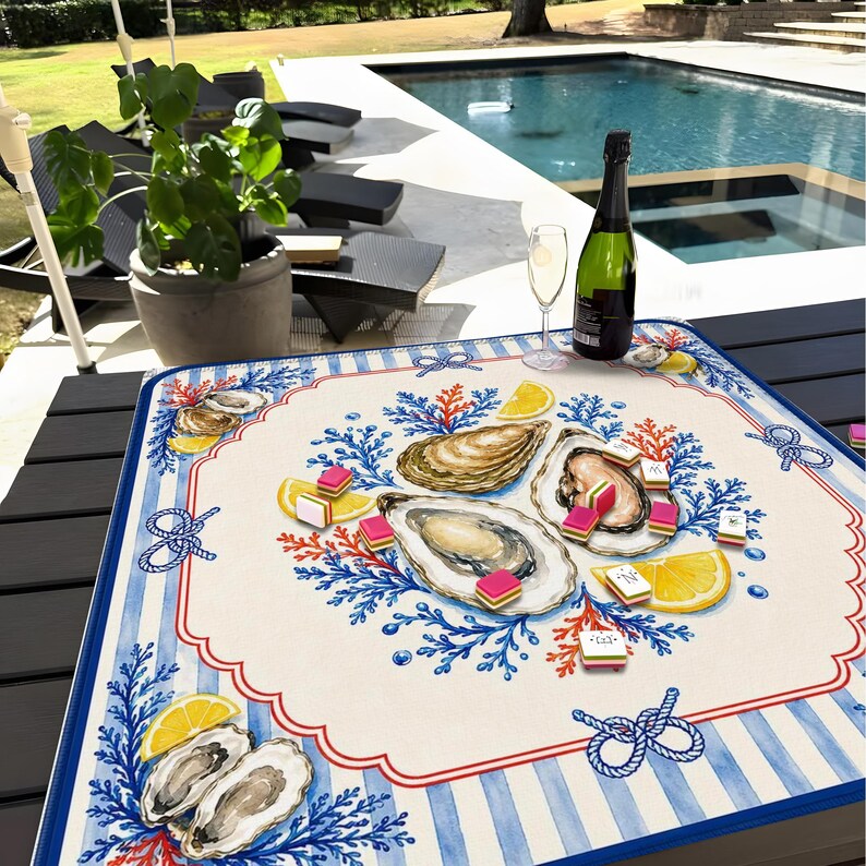 May include: A square, nautical-themed placemat with a cream-colored center featuring illustrations of oysters, lemon slices, and coral. The border has blue and white stripes, red trim, and rope knot designs. A bottle of champagne and a glass are on the table.