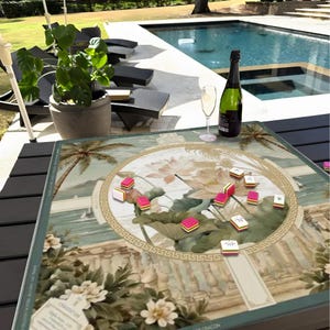 May include: A square game mat featuring a printed illustration of lotus flowers, palm trees, and classical architectural elements. The mat is scattered with small, rectangular game tiles in white, pink, and green, alongside a bottle and a glass.