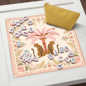 May include: A square mahjong mat featuring a cream background with a central illustration of two leopards under a pink palm tree, surrounded by floral borders. Scattered white mahjong tiles with various symbols and American flags accompany a yellow pouch.