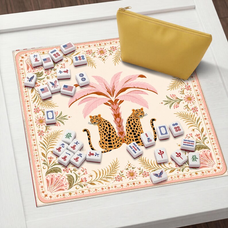 May include: A square mahjong mat featuring a cream background with a central illustration of two leopards under a pink palm tree, surrounded by floral borders. Scattered white mahjong tiles with various symbols and American flags accompany a yellow pouch.