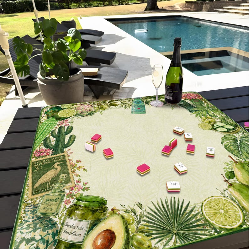 Puede incluir: Un tapete de juego cuadrado con un dise&ntilde;o bot&aacute;nico con aguacates, limas y hojas tropicales. Peque&ntilde;as piezas de juego coloridas est&aacute;n dispersas por el tapete. Una botella de champ&aacute;n y una copa est&aacute;n sobre el tapete.