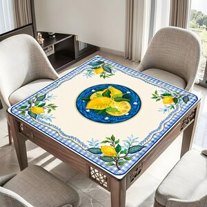 May include: Square table with a cream-colored tablecloth featuring a blue and white checkered border. The tablecloth has a central blue circle with a lemon illustration. Lemon and leaf motifs are also in the corners.
