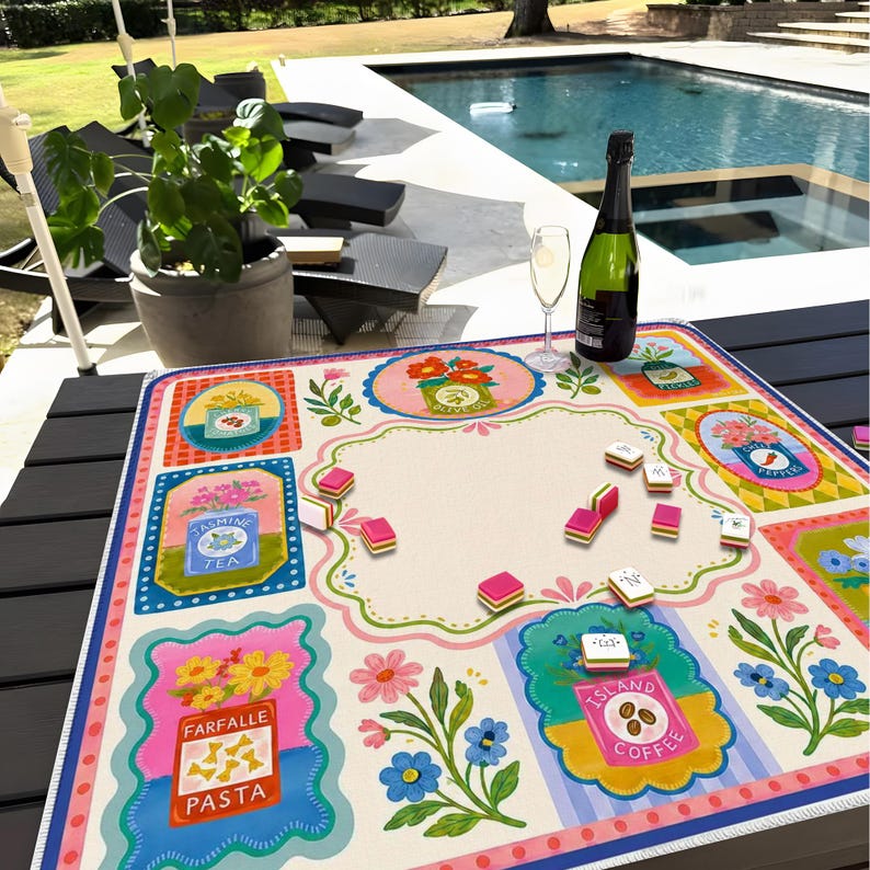 May include: A square game mat featuring a cream-colored center surrounded by colorful rectangular borders with illustrations of pantry items like Jasmine Tea, Farfalle Pasta, and Island Coffee. Small rectangular game tiles are scattered on the surface.