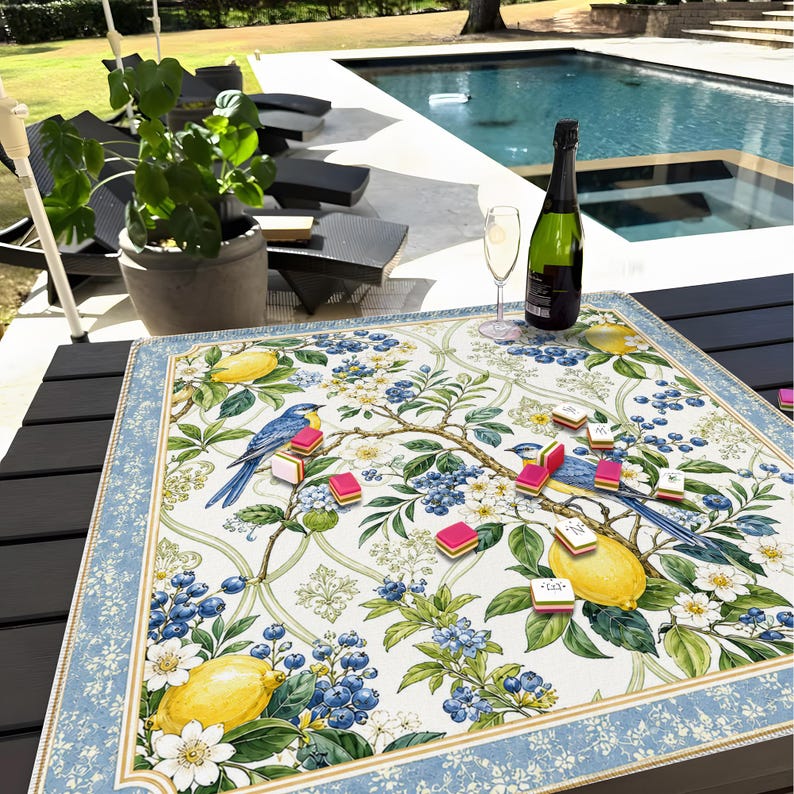 K&ouml;nnte beinhalten: Eine quadratische Tischdecke mit einem botanischen Druck aus gelben Zitronen, blauen Beeren, wei&szlig;en Bl&uuml;ten und blauen V&ouml;geln auf wei&szlig;em Grund, umrahmt von einer hellblauen Bord&uuml;re mit wei&szlig;em Blumenmuster, mit kleinen Spielsteinen darauf.