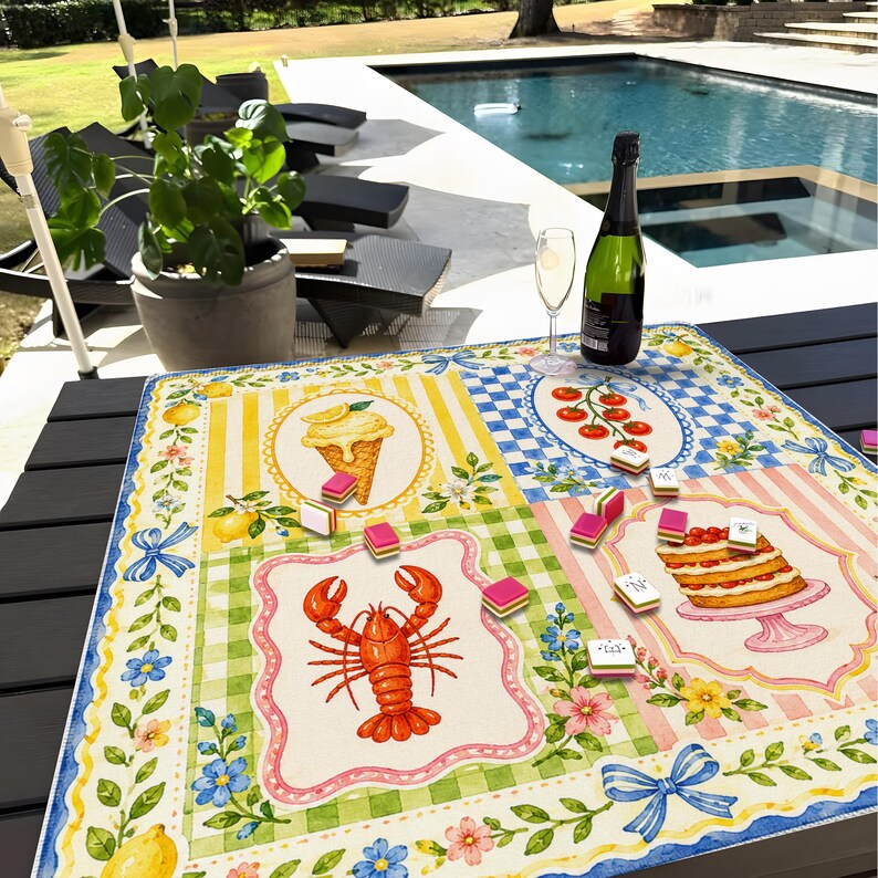 May include: A colorful, square tablecloth with a whimsical design featuring illustrations of a lobster, ice cream cone, cake, and tomatoes. The tablecloth has a border with lemons, flowers, and bows. A bottle of wine and a glass are on the table.