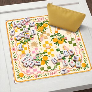 May include: A yellow zippered pouch rests on a decorative Mahjong mat with a floral and citrus fruit design. The mat features Mahjong tiles with various symbols and numbers, including American flag tiles. The scene is set on a white surface.