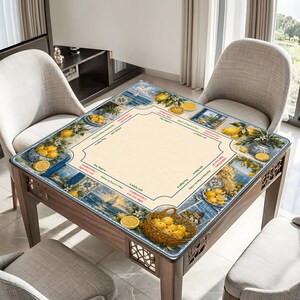 May include: A square wooden table with a decorative top featuring a lemon and coastal design. The table's surface has a central beige area surrounded by blue and white illustrations of lemons, vases, and seaside scenes. A woven basket of lemons sits on the table.