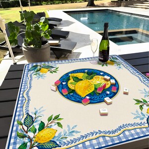 May include: A square, cream-colored tablecloth with a blue and white gingham border and lemon illustrations. A blue plate in the center features lemon slices. Several game pieces are scattered on the table, along with a bottle of champagne and a wine glass.