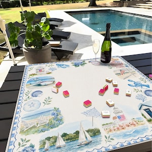 May include: A square fabric game mat featuring a coastal watercolor print with lighthouses, sailboats, beach chairs, and seashells. Scattered across the white center are several small, rectangular, multi-colored game tiles with pink edges.