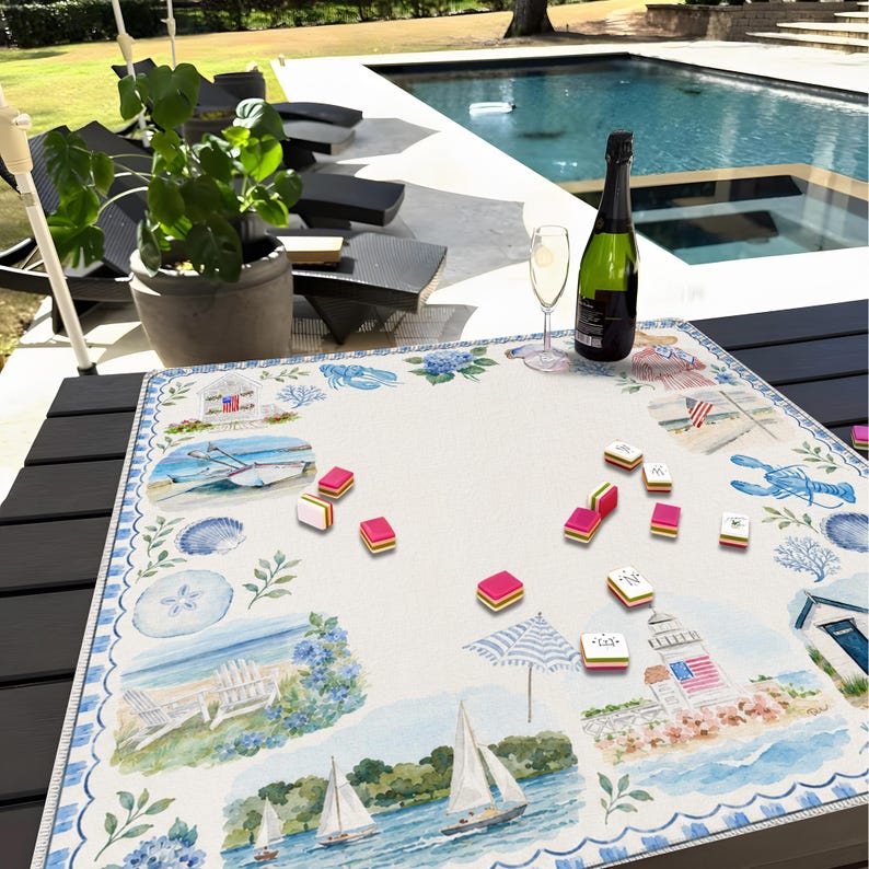May include: A square fabric game mat featuring a coastal watercolor print with lighthouses, sailboats, beach chairs, and seashells. Scattered across the white center are several small, rectangular, multi-colored game tiles with pink edges.