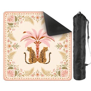 May include: A square outdoor blanket featuring a cream background with a central pink palm tree and two leopards. The border has floral and fern motifs in pink, green, and orange. The blanket has a black underside and includes a black carrying bag.
