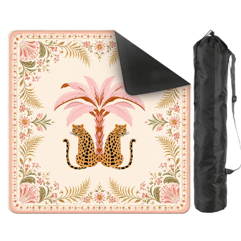 May include: A square outdoor blanket featuring a cream background with a central pink palm tree and two leopards. The border has floral and fern motifs in pink, green, and orange. The blanket has a black underside and includes a black carrying bag.