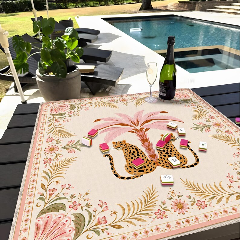 May include: A square game mat featuring a central illustration of two leopards sitting under a pink palm tree, surrounded by a floral border in shades of pink, green, and gold. Several small rectangular game tiles are scattered across the surface.