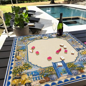 May include: A decorative game board featuring a coastal Italian scene with a yellow scooter, lemons, and blue accents. Pink and white game tiles are scattered on the beige playing surface. A bottle of wine and a champagne glass are on the table.