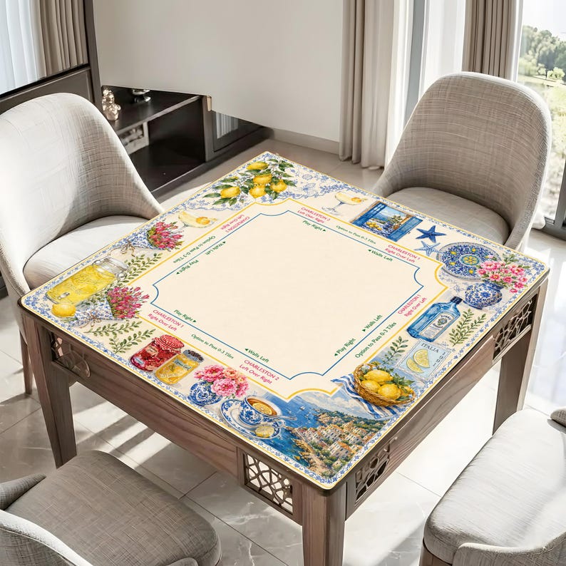May include: A square wooden table with a decorative top featuring a lemon and floral design. The table has a light beige center with a blue border and illustrations of lemons, flowers, and Italian scenes. The table is surrounded by two gray chairs.