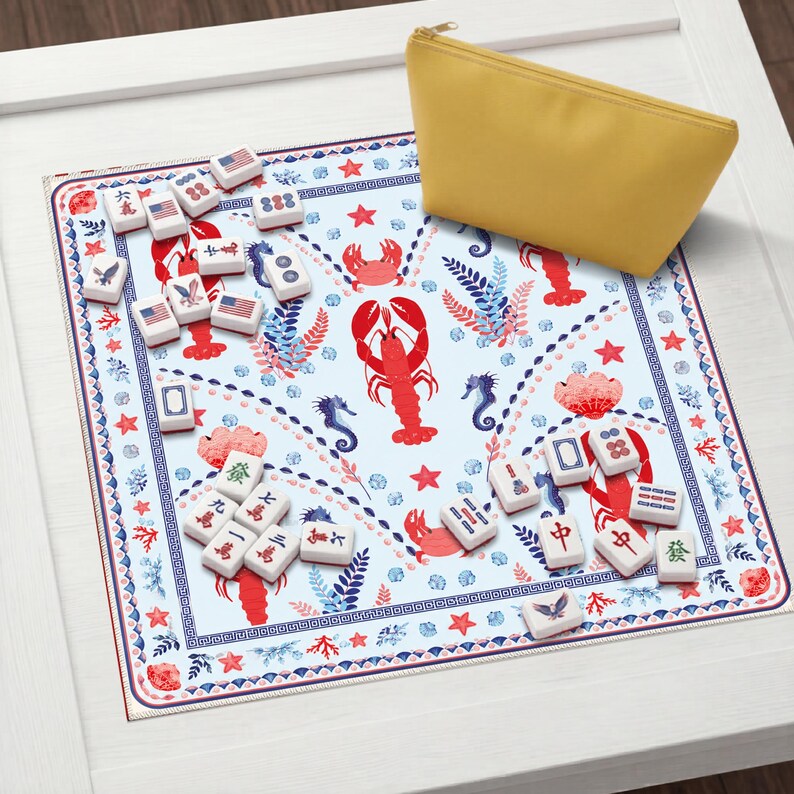K&ouml;nnte beinhalten: Eine quadratische Spielmatte mit hellblauem Hintergrund, verziert mit einem Muster aus roten Hummern, Krabben und Seepferdchen, umrandet von einem blau-roten geometrischen Design. Wei&szlig;e Mahjong-Steine mit Symbolen und US-Flaggen liegen neben einer gelben Tasche.