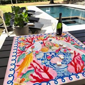 May include: A colorful square game mat featuring two white cockatoos, coral, and a blue vase. The mat has a pink border with a blue wave design. A bottle of champagne and a glass are on the table.